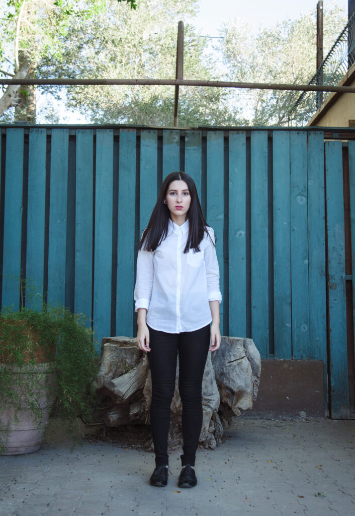 Gabriela Inzunza stands against a teal wooden backdrop wearing a white shirt and black pants with a rustic wooden bench and green plants in the foreground.